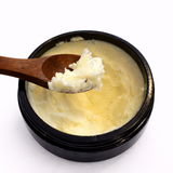 Black container with yellowish substance (Athena's Chrism: Deep Moisturizer) and wooden spoon on a white background