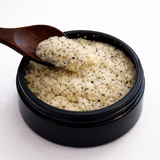Black container of salt (Bright Dot's Body Scrub) with a wooden spoon on a white background