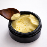 Black container with yellow cream (Catalyst Massage Mousse) and a wooden spoon on a white background