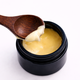 Wooden spoon lifting a scoop of yellow cream (Esha's Eye Cream) from a black container on a white background