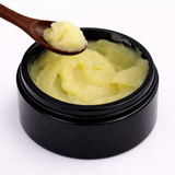 Black container with yellowish substance (Methuselah's Magik Breast Balm) and a wooden spoon on a white background