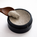 Black container with beige powder (Pollen Grains: Face Scrub) and a wooden spoon on a white background