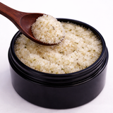 Black container with a wooden spoon holding a scoop of white powder (The Man Scrub: Soothes Hardworking Skin) on a white background